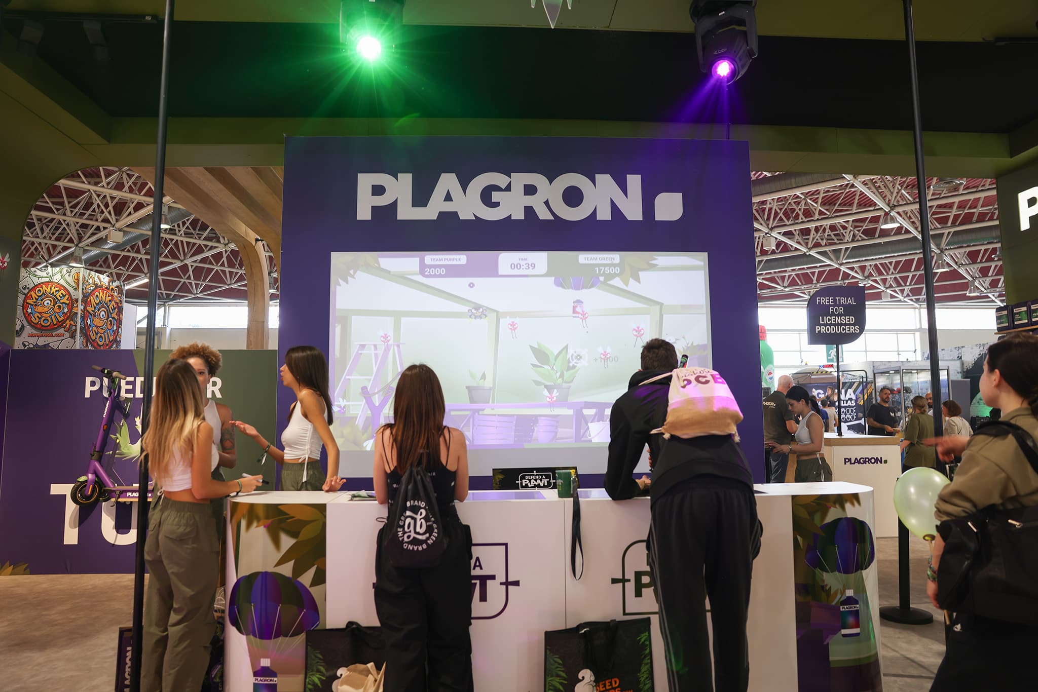 The Plagron booth setup at a trade show. Two visitors play side by side on the big screen while Team Purple and Team Green scores tick up live.